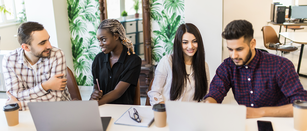 A diverse group of four people are sitting around a table, working on laptops