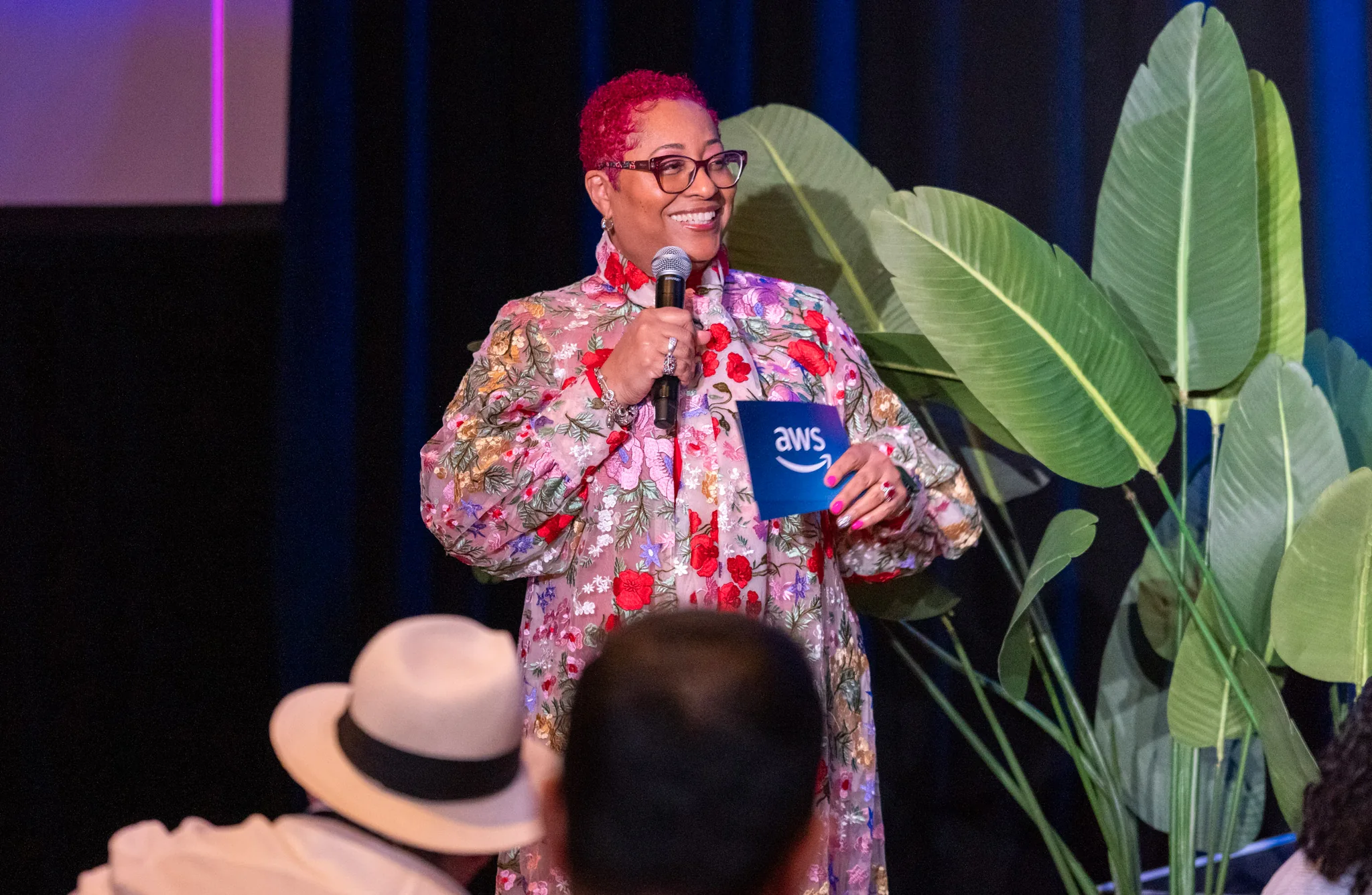 Speaker in a floral jacket holding a microphone while addressing an audience.