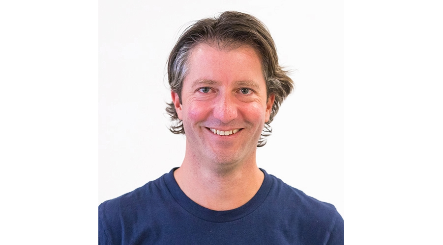 A person with wavy brown hair wearing a navy blue shirt against a plain white background.