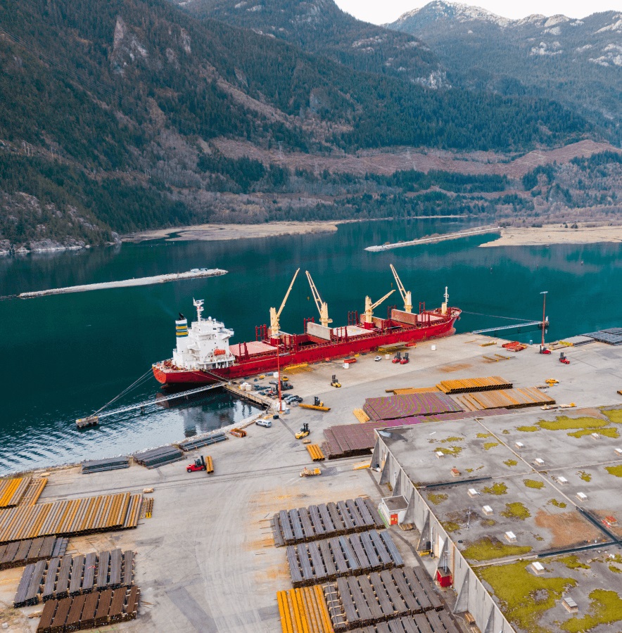 A red cargo ship docked at an industrial port surrounded by stacked materials, with a calm lake and forested mountains in the background.