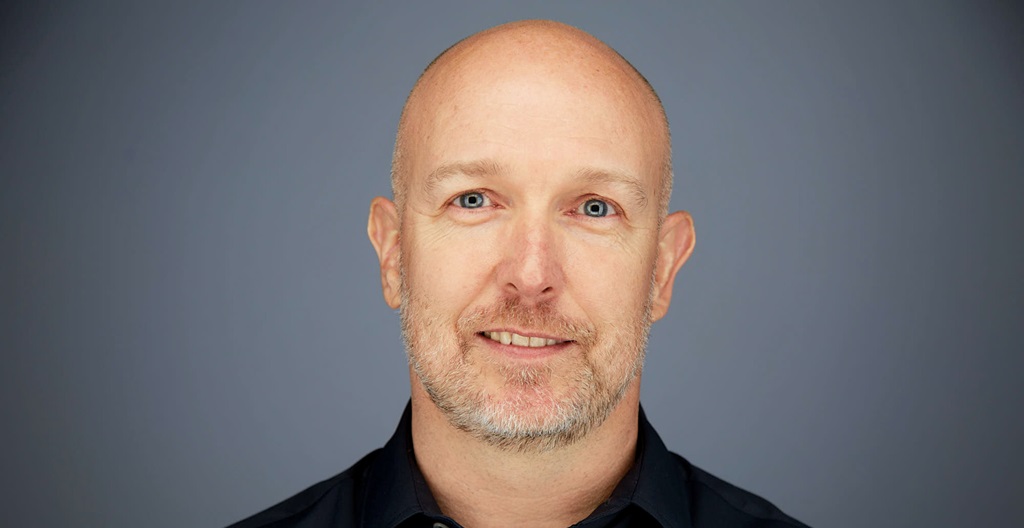 A person wearing a dark shirt against a plain gray background.
