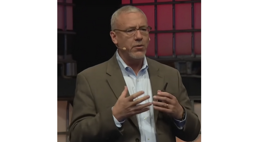 A person in a brown blazer and light blue shirt gesturing with their hands while speaking on stage with a red and black background.