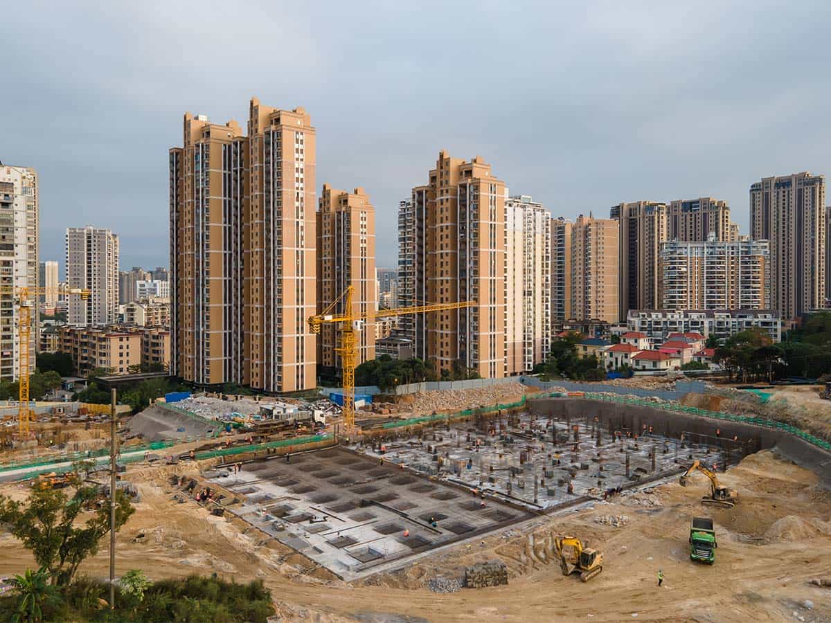 Construction site with heavy machinery in the foreground, surrounded by tall beige apartment buildings in an urban area.