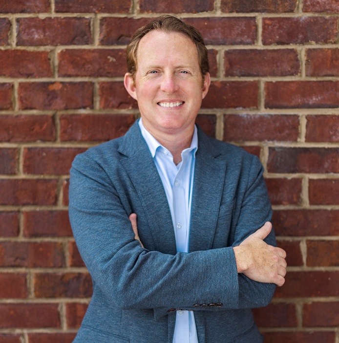 A person in a blue blazer and light blue shirt standing against a red brick wall with arms crossed.