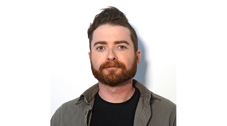 A person wearing a black shirt and an olive-green jacket against a white background.