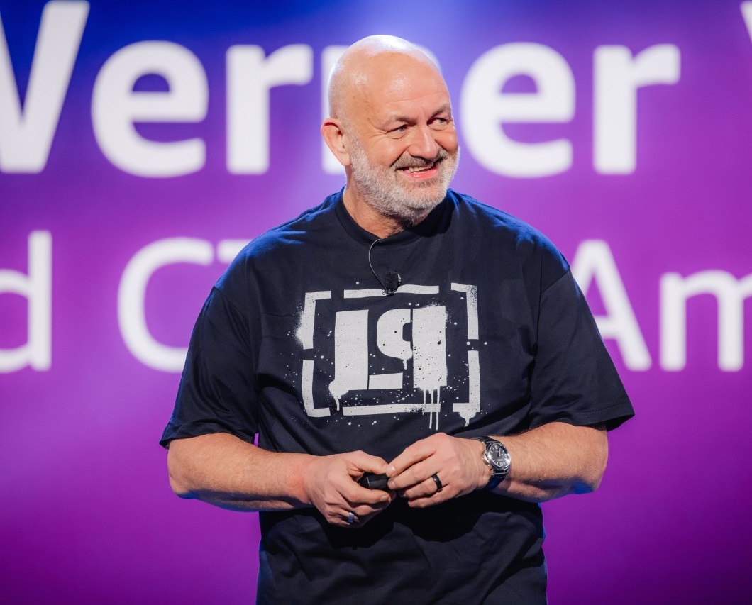 A person wearing a black graphic t-shirt with a microphone headset, standing in front of a purple background with partially visible white text.