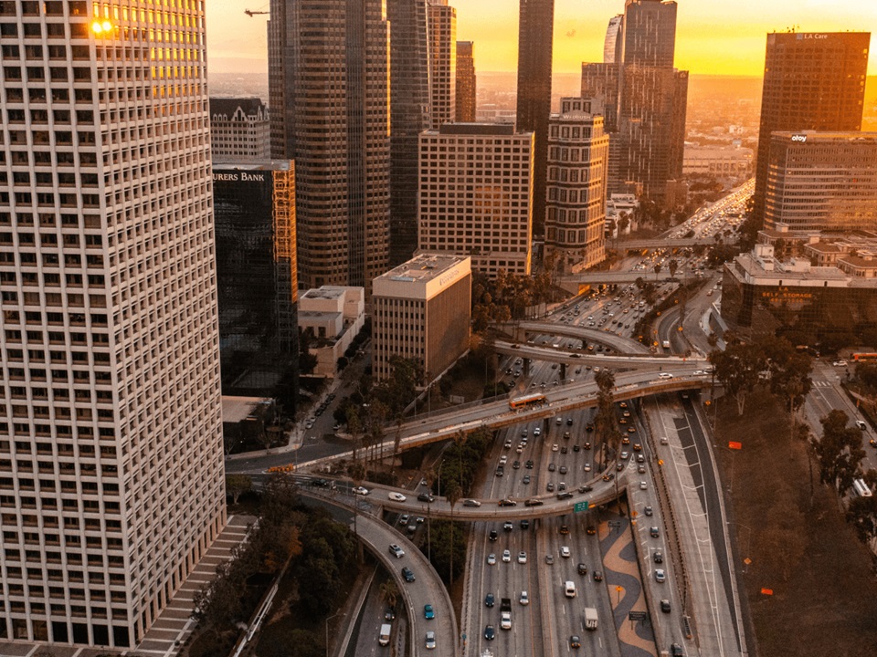 Aerial view of a busy freeway interchange surrounded by tall skyscrapers in downtown Los Angeles at sunset.