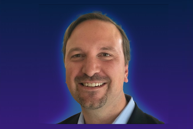 A person wearing a suit jacket and collared shirt against a gradient blue background.