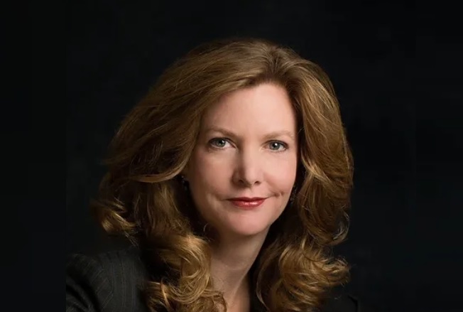 Alt-text: Woman with wavy auburn hair wearing a dark blazer against a black background.