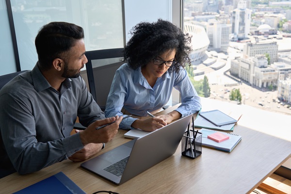 Two professionals collaborating at a desk in a modern office environment with a laptop, notebooks, and city view in the background.