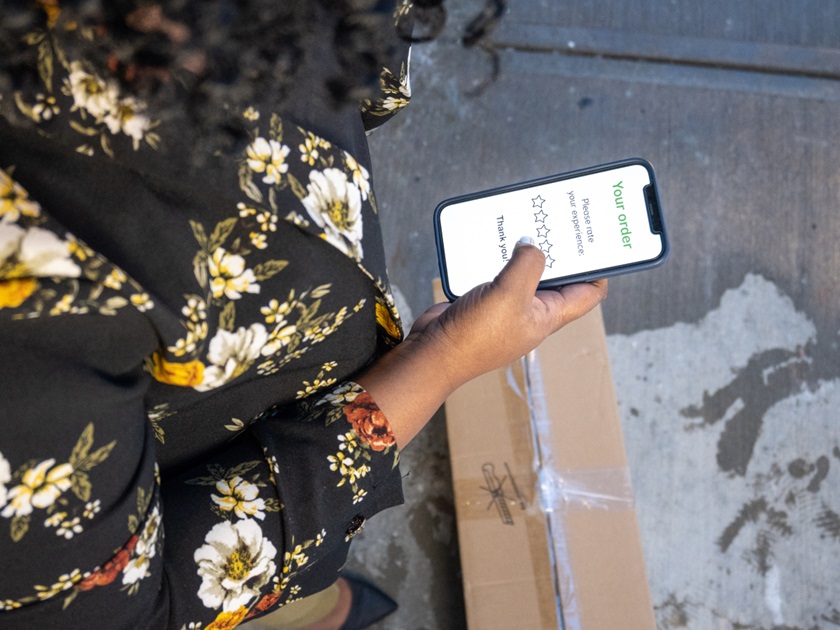 A person holding a smartphone displaying a delivery rating screen, standing next to a cardboard package on a concrete surface.