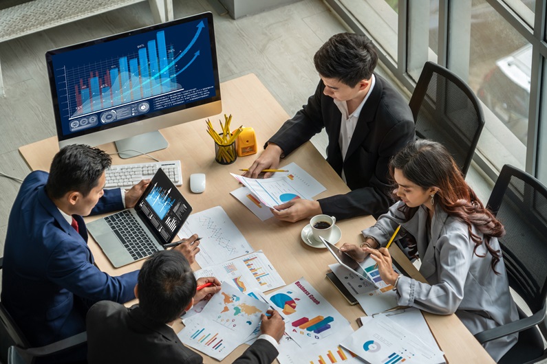 A group of professionals in a meeting analyzing charts and graphs on paper, a tablet, and a computer screen displaying data visualizations.