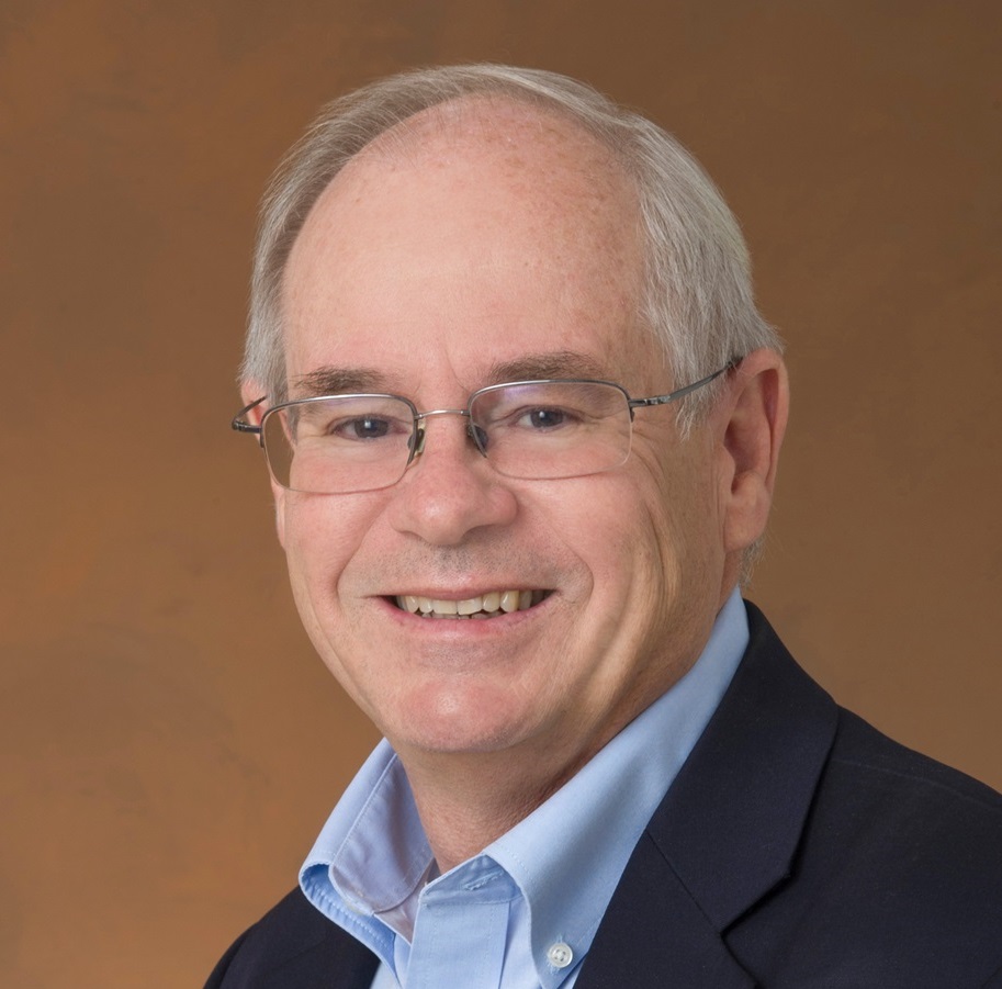 A person wearing a blue collared shirt and dark blazer against a brown background.