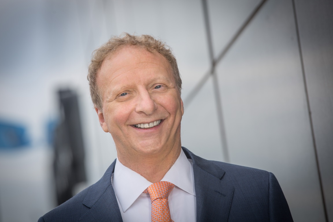 A person wearing a navy suit, white shirt, and orange tie, standing in front of a modern glass building.