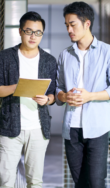 Two people standing indoors, one holding a folder and the other with hands clasped.
