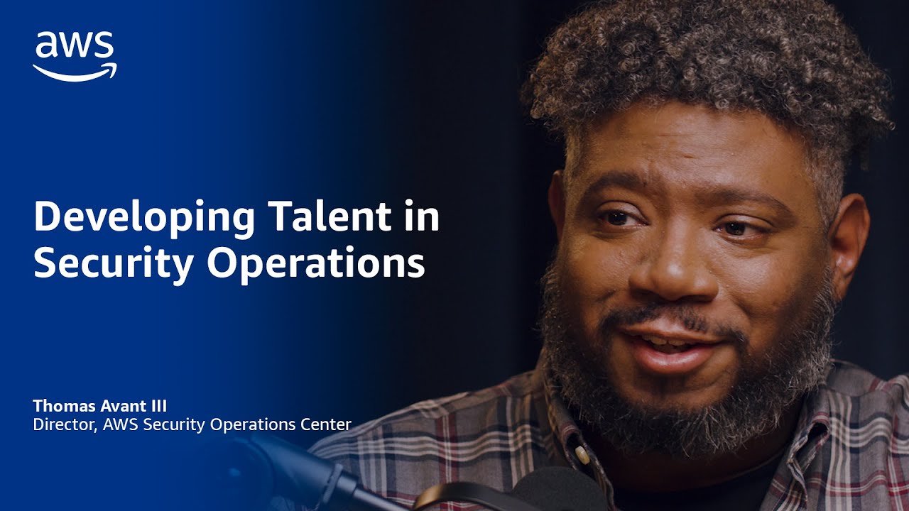 Text on a blue background reads "Developing Talent in Security Operations," with "Thomas Avant III, Director, AWS Security Operations Center" below. A microphone and plaid shirt are partially visible.