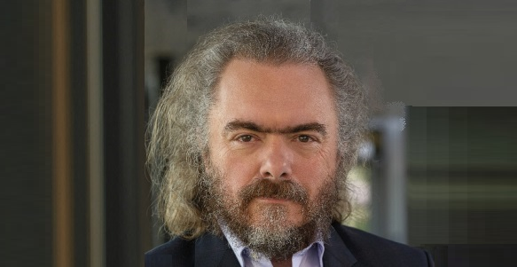 A person with curly, gray hair wearing a dark suit and white shirt, standing indoors near a window.
