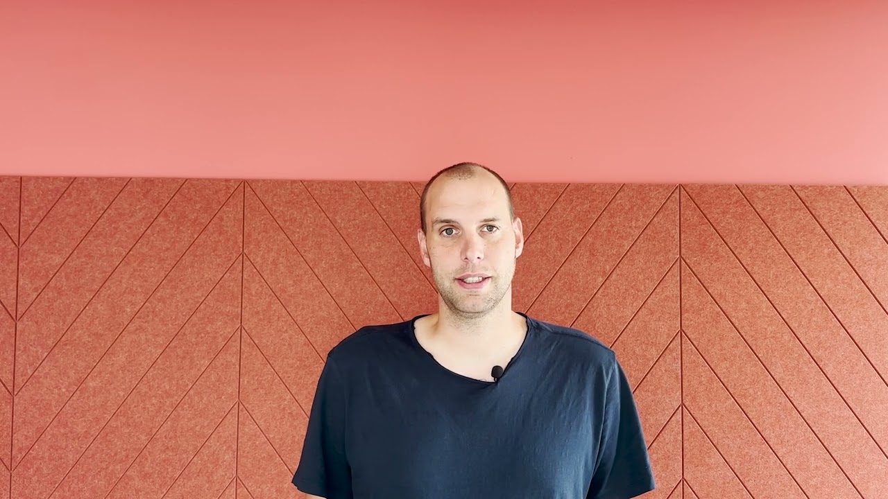 A man wearing a black shirt stands in front of a red geometric-pattern wall. He is facing the camera and the background features a textured design with chevron patterns.