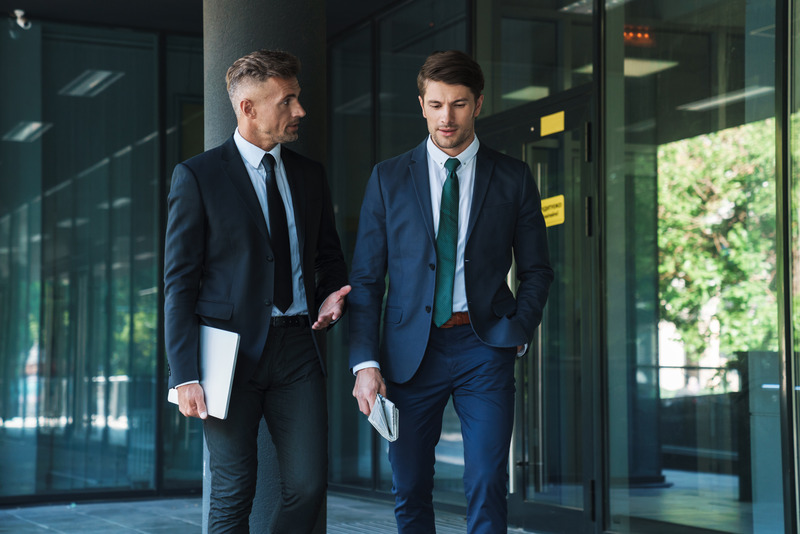 Two business professionals in suits walking and talking outside a modern office building. One holds a laptop and the other carries a newspaper.