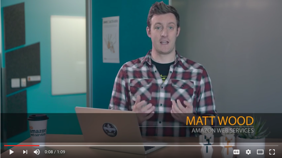 A screenshot from a video presentation featuring Matt Wood from Amazon Web Services discussing AWS Lifecycle Hooks. Matt is seated at a desk with an AWS-branded coffee cup and laptop, speaking to the camera in a modern office setting.