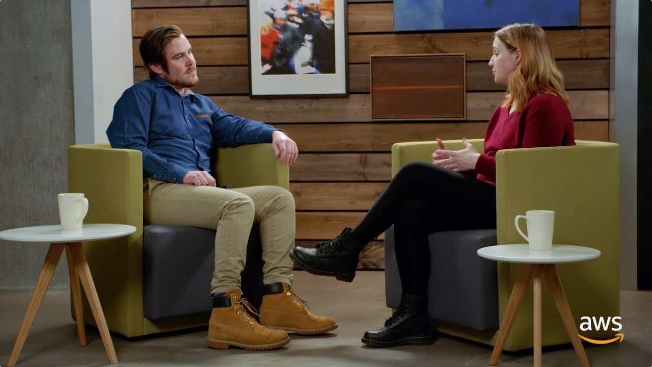 Two people having a conversation while seated in modern armchairs in an office setting with wooden walls, small side tables with coffee mugs, and AWS branding visible. This image is designed as a YouTube thumbnail.