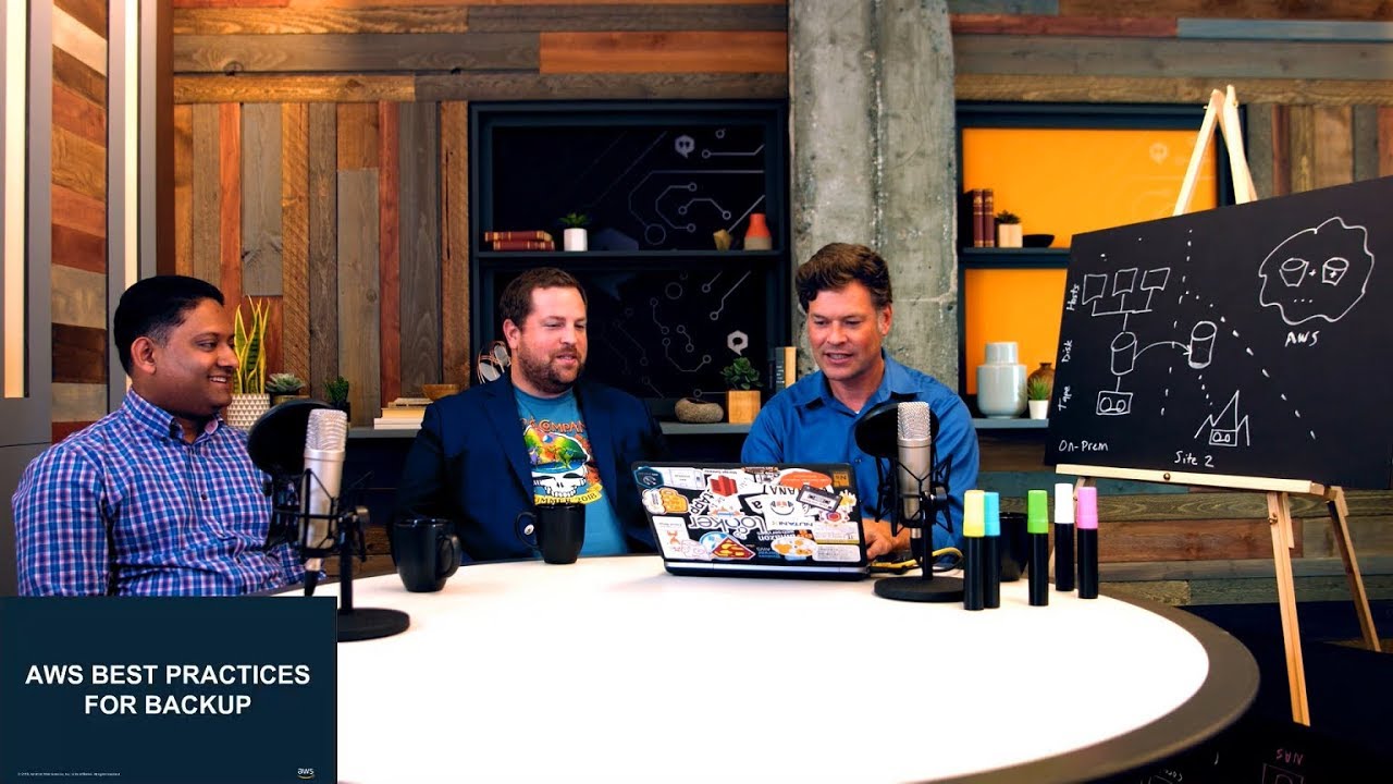 YouTube video thumbnail showing three presenters discussing AWS best practices for backup. They are sitting at a round table with microphones and a laptop, with a chalkboard diagram explaining backup strategies visible in the background.