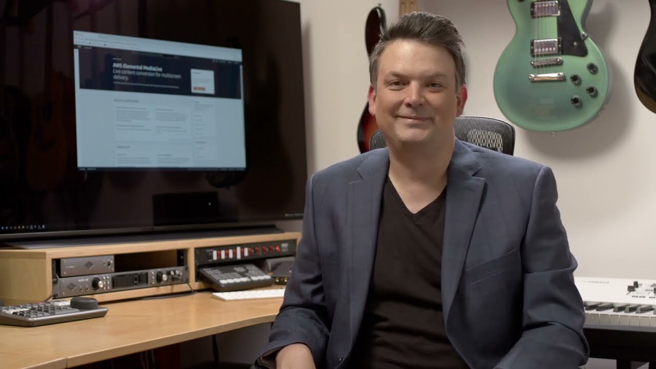 A man sits in a music studio with various guitars on the wall, music equipment, and a large computer screen displaying an AWS webpage.