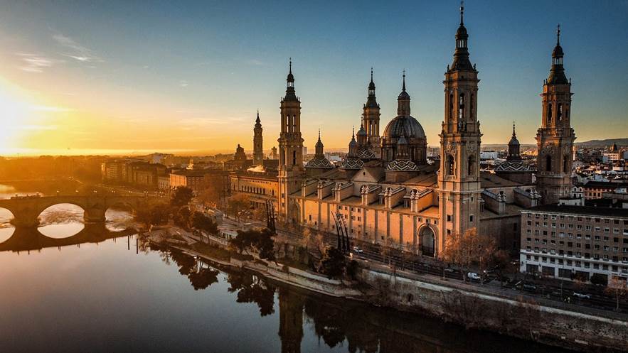 A stunning sunrise view of the Basilica del Pilar, a historic cathedral on the banks of the Ebro River in Zaragoza, Spain, with cityscape and warm morning light.