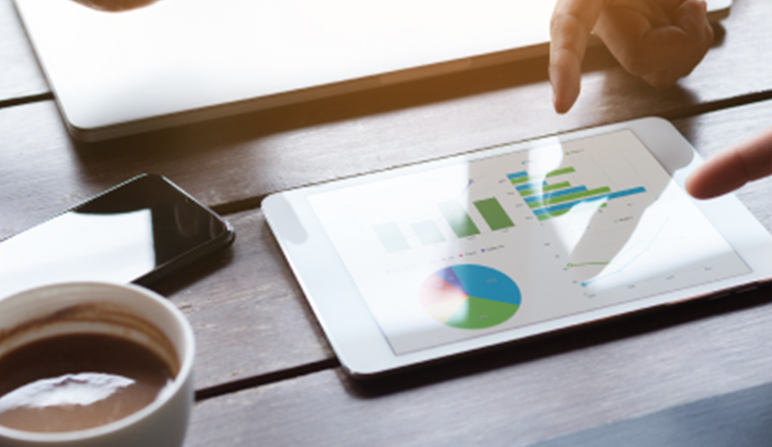 A tablet on a wooden table displays various charts and graphs while hands point at the screen. Also visible are a smartphone, a laptop, and a cup of coffee, suggesting a business or analytics discussion setting.