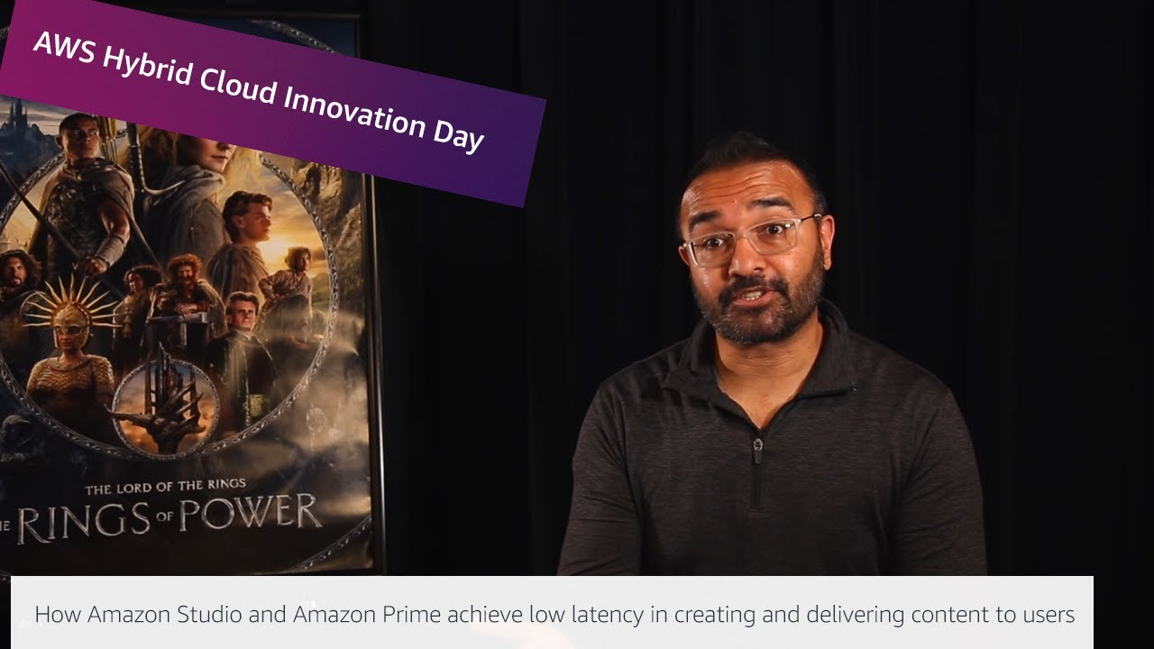 YouTube thumbnail featuring a speaker discussing how Amazon Studio and Amazon Prime achieve low latency in creating and delivering content to users, with a poster of 'The Lord of the Rings: The Rings of Power' in the background and a banner reading 'AWS Hybrid Cloud Innovation Day'.