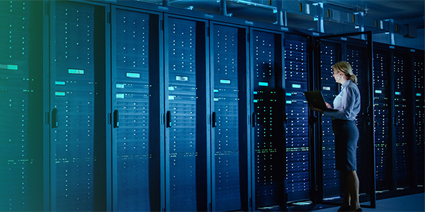 A person using a laptop stands in a server room filled with rows of data center racks, representing AWS global infrastructure.