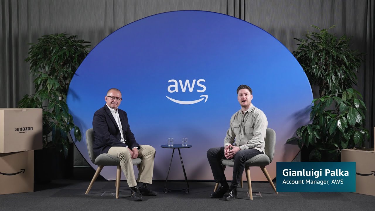 Two people seated in front of an AWS-branded backdrop having an interview, with plants and Amazon boxes in the background. The lower third identifies one guest as Gianluigi Palka, Account Manager, AWS.
