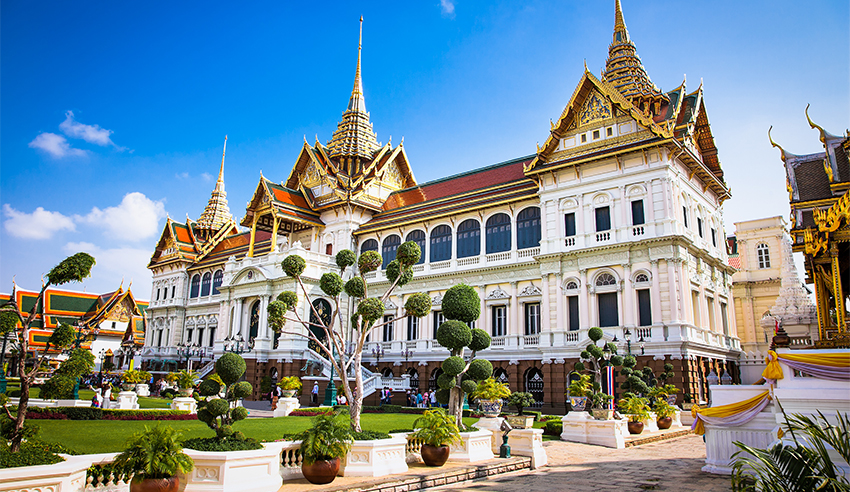 A garden view of the Grand Palace in Bangkok, Thailand, featuring ornate traditional Thai architecture and manicured trees under a clear blue sky.