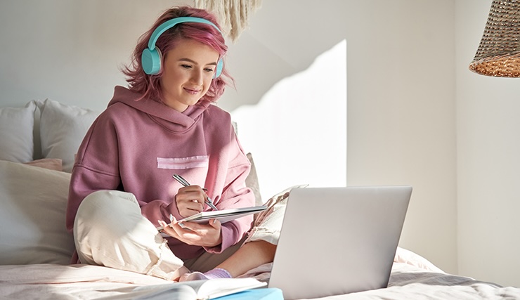 Hipster teen girl school student with pink hair wear headphone write notes watch video online webinar learn on laptop sit in bed distance elearning course video conference pc call in bedroom at home.