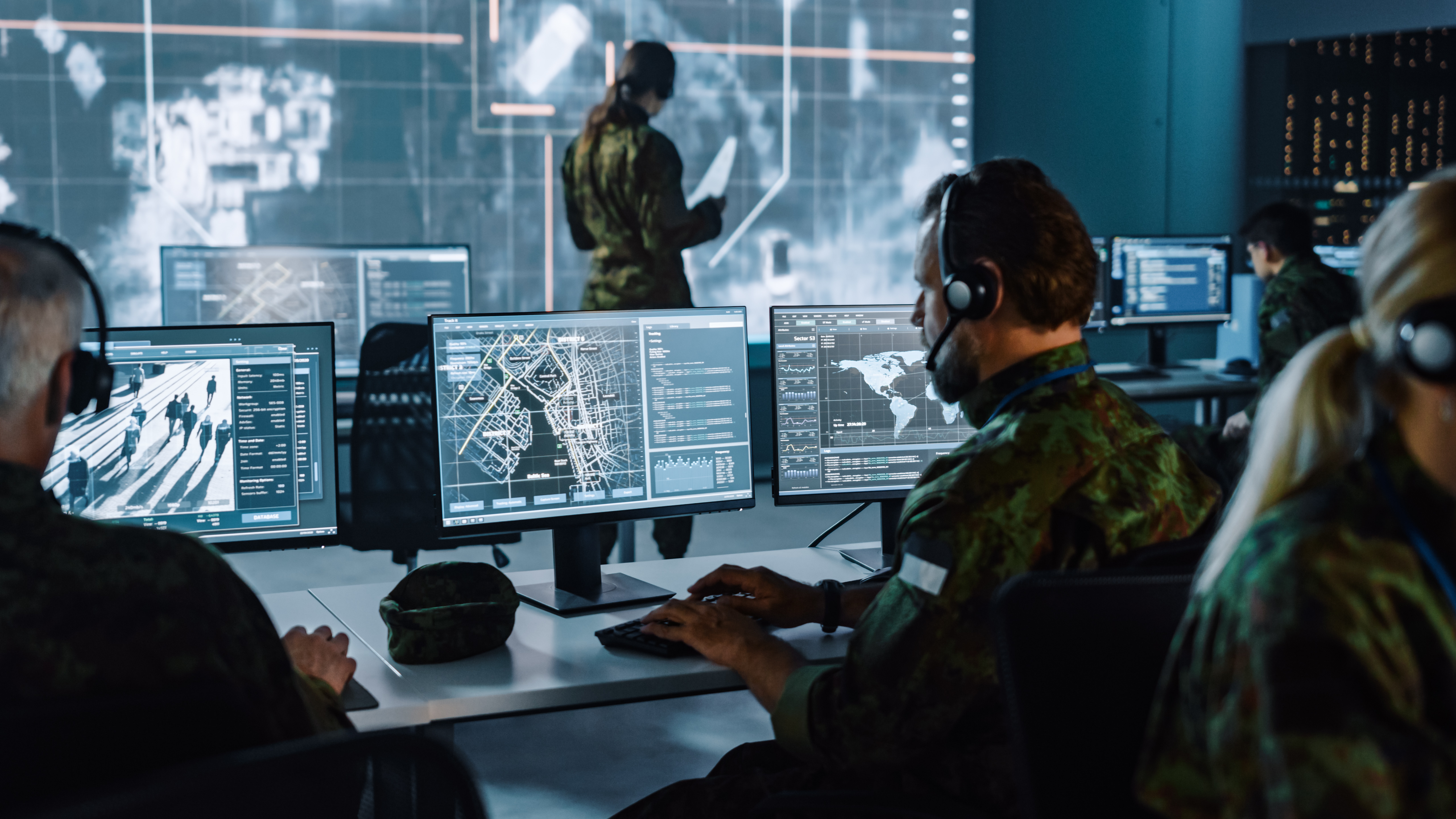 A team of uniformed personnel works inside a modern command and control center, illuminated by the cool glow of multiple digital displays. Seated at a row of desks, operators wearing camouflage uniforms and headsets monitor large computer screens filled with detailed city maps, live surveillance feeds, data charts, and global tracking interfaces.