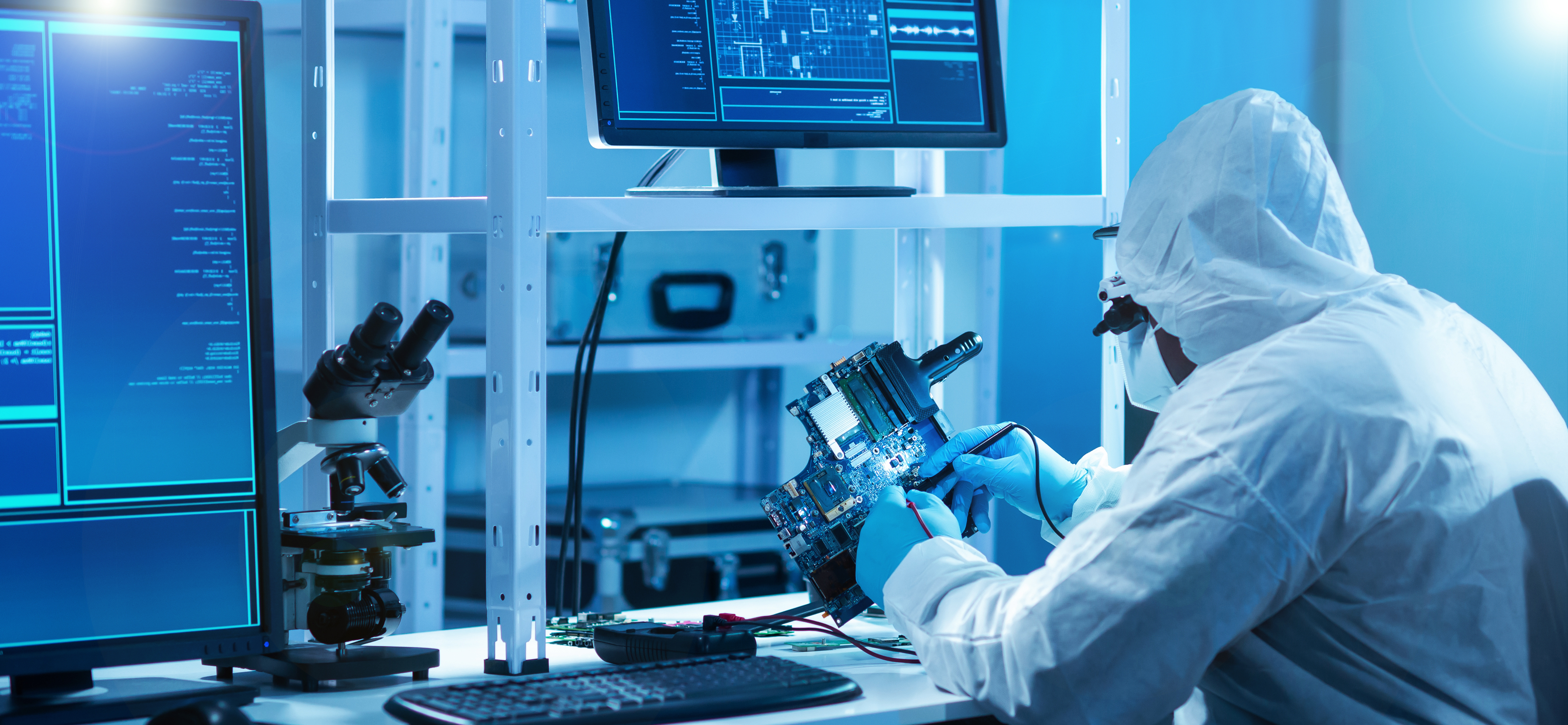 A technician in full protective gear works carefully inside a high-tech laboratory environment. Dressed in a white hooded cleanroom suit, gloves, and protective eyewear, the individual examines a complex circuit board under bright, cool-toned lighting.