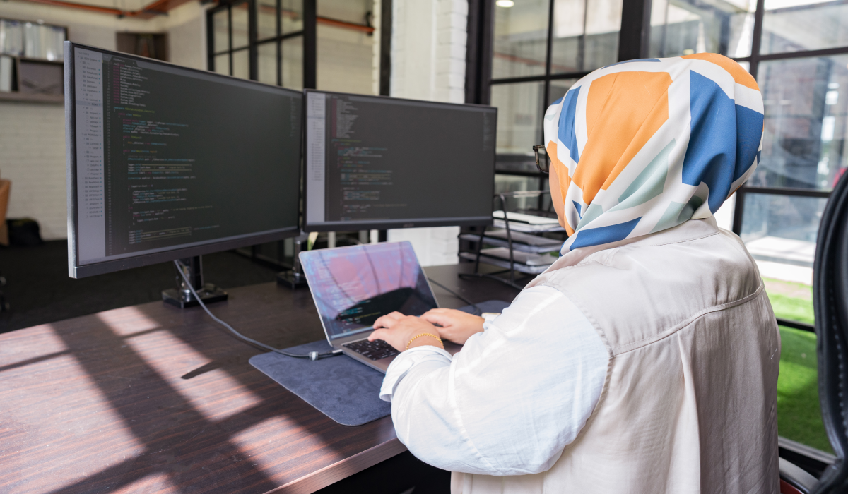 A woman wearing a colorful headscarf works on a laptop and two large monitors displaying code in a modern office environment.