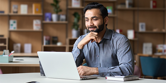 A man sits at a desk in a modern office space, looking at a laptop and smiling, with books and shelves in the background.