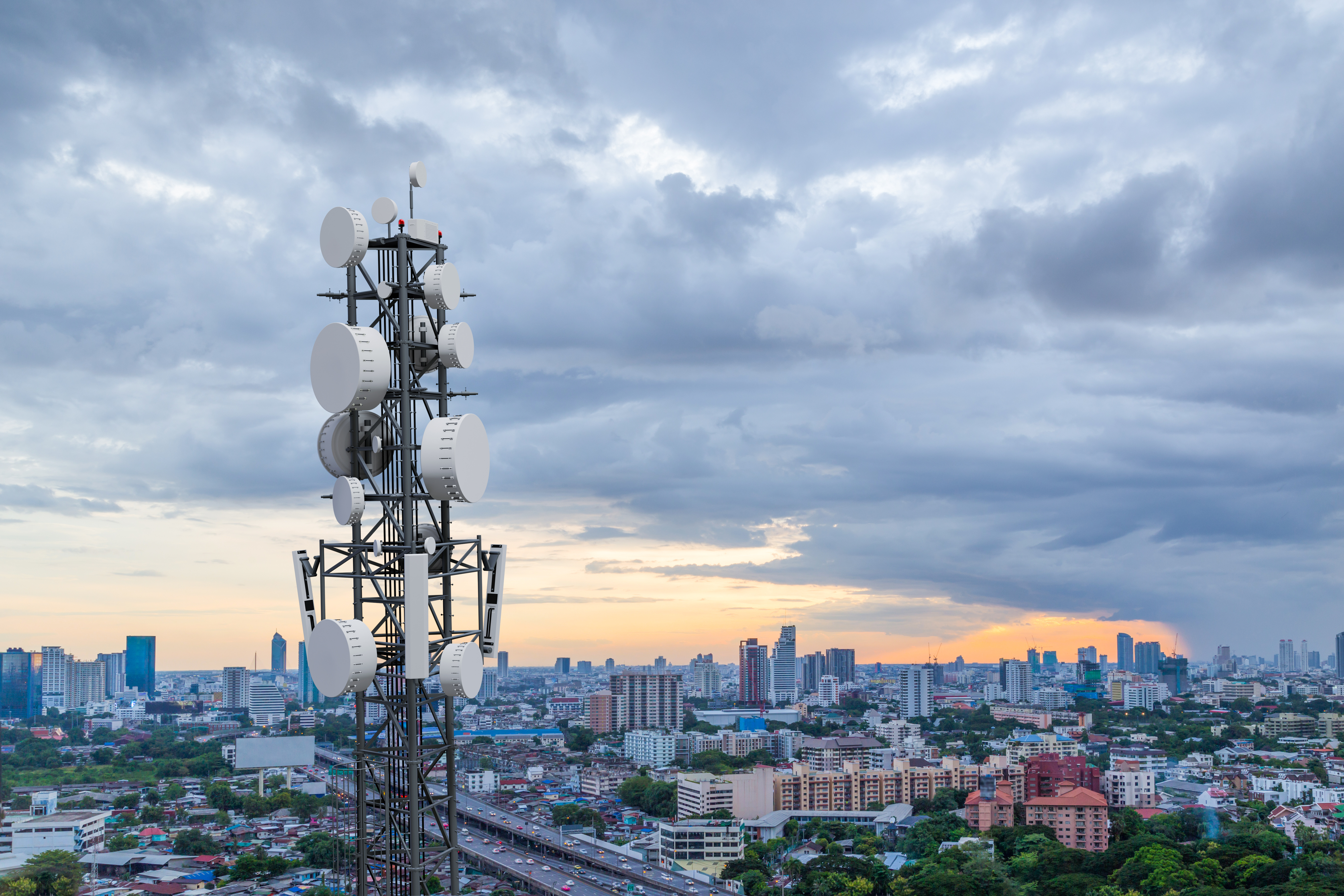 A 5G telecommunication tower stands tall in the foreground with multiple antennas, overlooking a modern cityscape at sunset under a partly cloudy sky.