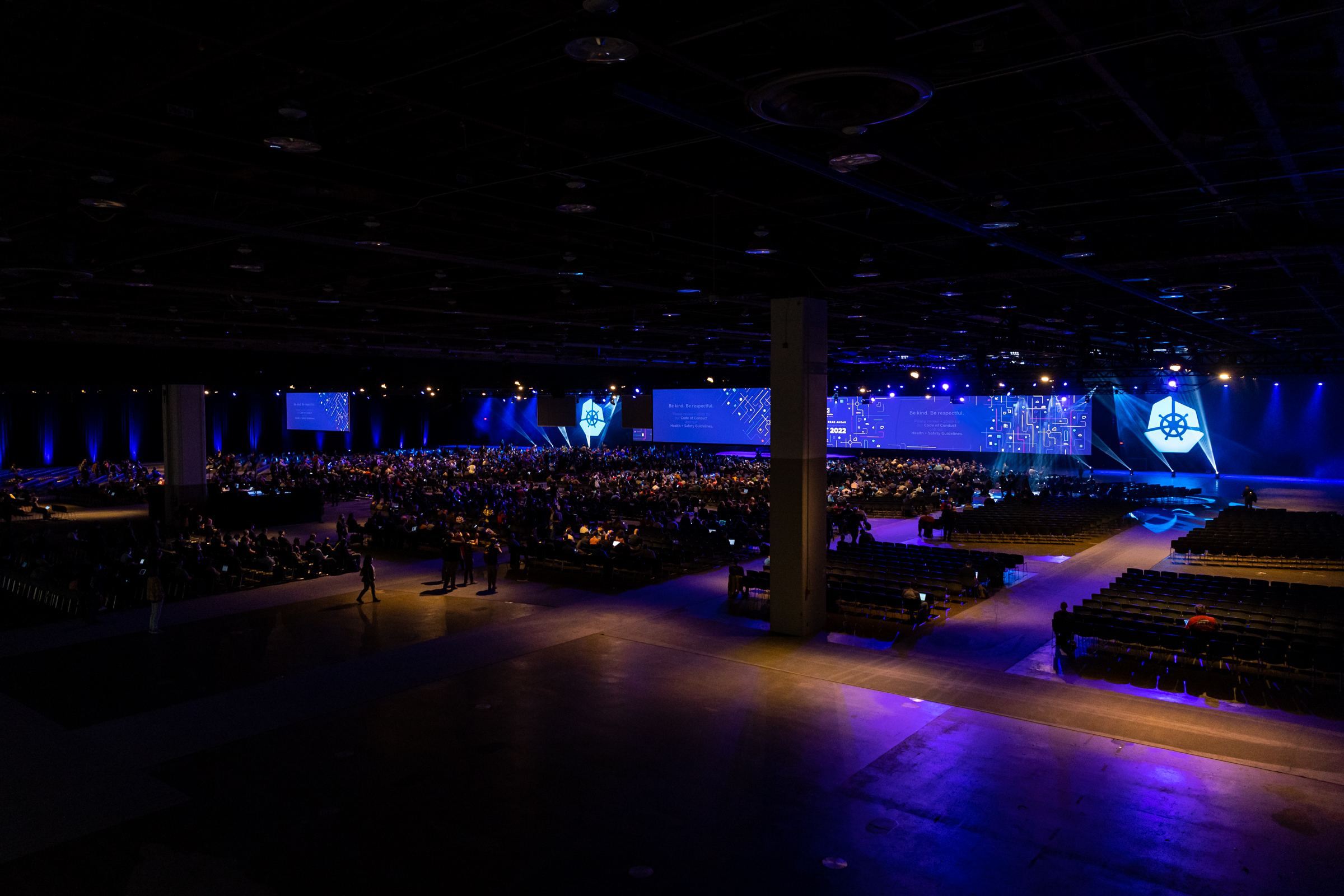 Large audience at an AWS-sponsored KubeCon event in a convention hall, with prominent Kubernetes branding and blue stage lighting.