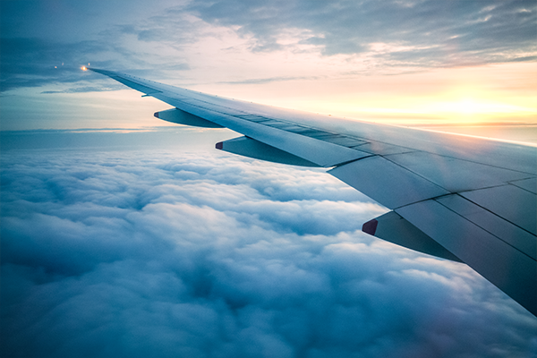 A view of an airplane wing above a blanket of clouds during sunset, with warm sunlight on the horizon.