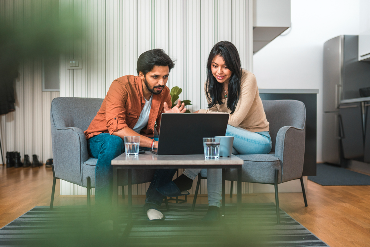 A couple sitting together on a sofa in a modern living room, working on a laptop placed on a coffee table. Both appear focused and engaged in their work, with glasses of water nearby.