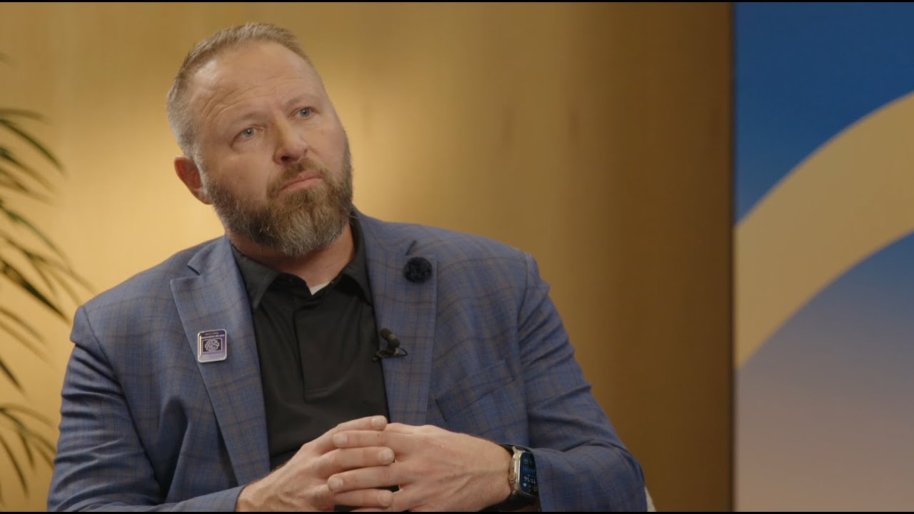 A man with a beard, wearing a blue suit and black shirt, sits for an interview. He appears thoughtful, with hands clasped, against a warm-toned background. This image is used as a YouTube video thumbnail.