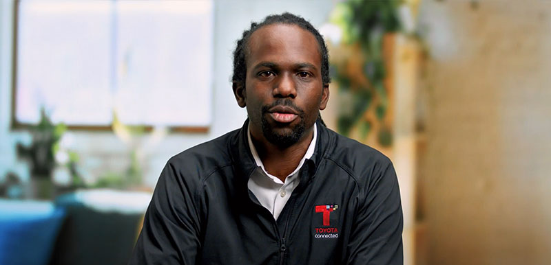 A Toyota Connected employee wearing a branded jacket is pictured in a professional indoor setting with a soft focus background.