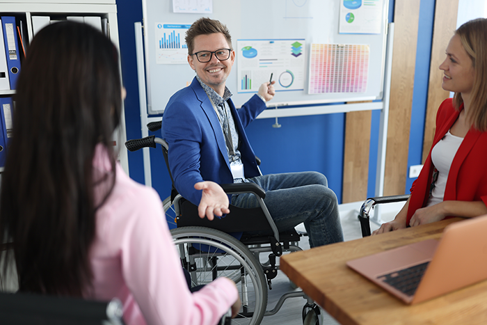 A man in a wheelchair wearing a blue blazer gestures toward a whiteboard with charts, while two women listen during a meeting.