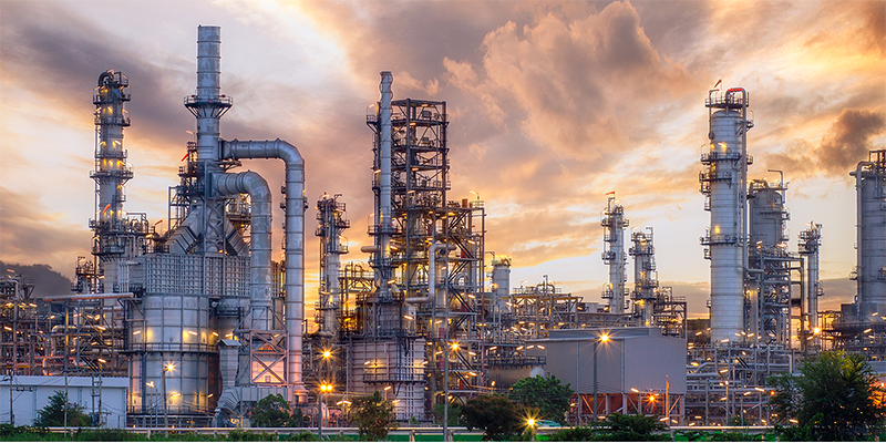 Industrial refinery complex with tall metal structures and chimneys, illuminated by lights at sunset under a cloudy sky.