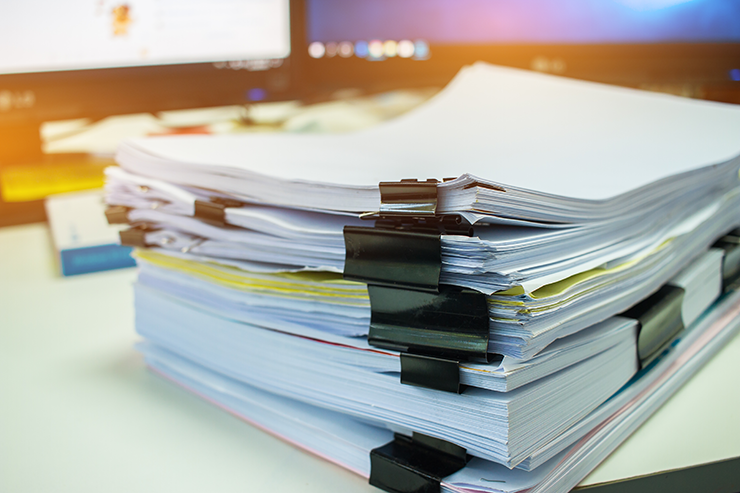 A stack of paper documents bound with large binder clips placed on a desk, with two computer monitors in the background.