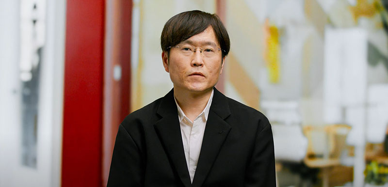 A portrait of a Honda executive wearing glasses and a black suit, seated in a modern office environment with a blurred background.