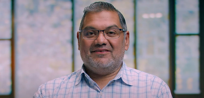 A man with glasses and a checkered shirt is smiling, standing in front of blurred large windows in the background.