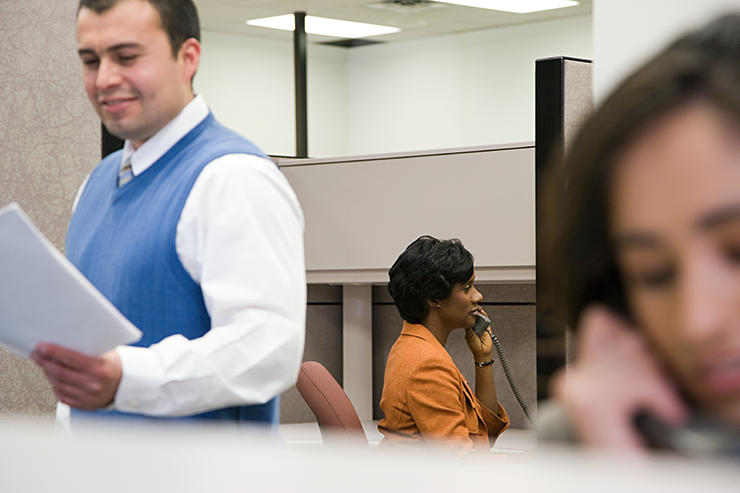 A woman in an orange blazer sits at a desk in a cubicle, speaking on the phone, with an office setting in the background.